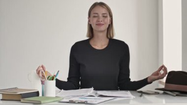 Sakin, genç, sarışın bir kadın meditasyon pozunda parmaklarını tutarak beyaz bir ofiste masanın içinde oturuyor.