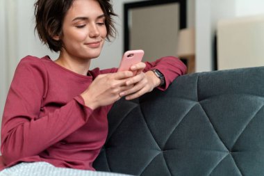 Oturma odasındaki kanepede otururken akıllı telefon kullanan rahat elbiseli genç bir kadının fotoğrafı.