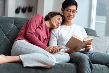 Çok uluslu çiftin kitap okuduğu ve evdeki koltukta otururken akıllı telefon kullandığı bir fotoğraf.