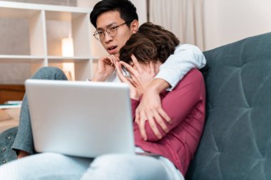 Korkmuş çokuluslu çiftin evdeki kanepede otururken birbirlerine sarılıp dizüstü bilgisayar kullanmalarının fotoğrafı.