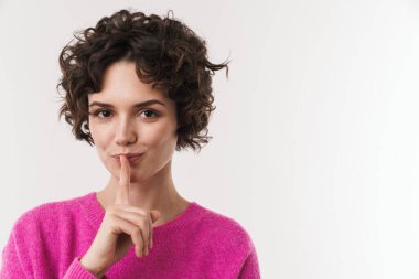 Memnun kıvırcık kadın, sessiz bir jest yapıyor ve beyaz arka planda izole edilmiş kameraya bakıyor.