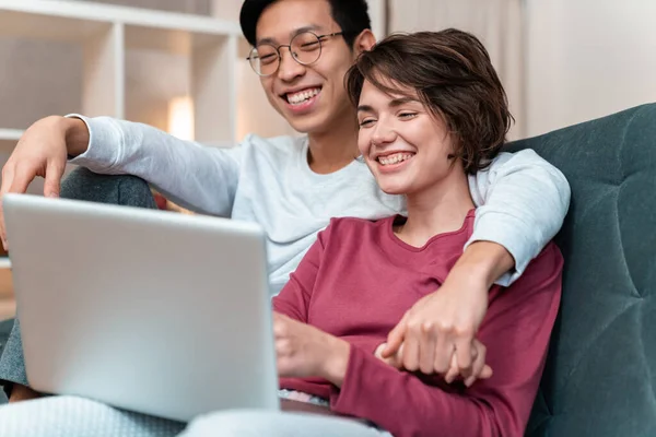 Foto Alegre Pareja Multinacional Riendo Usando Portátil Mientras Está Sentada —  Fotos de Stock