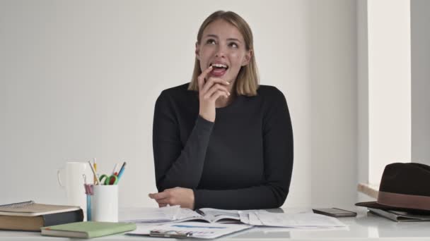 一位金发碧眼的年轻女子笑着梦想着在屋里的一个白色办公室里的桌子旁坐着什么东西 — 图库视频影像