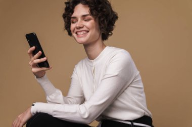 Bej arka planda tek başına otururken cep telefonu kullanan gülen güzel bir kadın görüntüsü