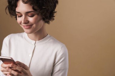 Neşeli, baştan çıkarıcı bir kadının bej arka planda izole edilmiş bir şekilde cep telefonu ve gülümsemesi.
