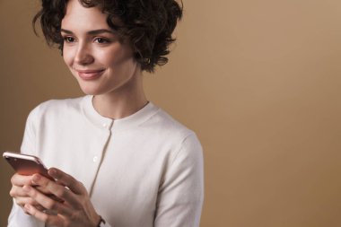 Neşeli, baştan çıkarıcı bir kadının bej arka planda izole edilmiş bir şekilde cep telefonu ve gülümsemesi.