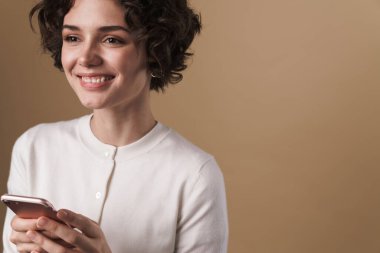 Neşeli, baştan çıkarıcı bir kadının bej arka planda izole edilmiş bir şekilde cep telefonu ve gülümsemesi.