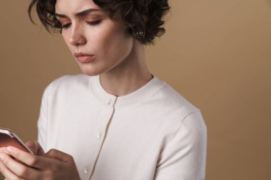 Bej arka planda izole edilmiş cep telefonu kullanan ve düşünen ciddi baştan çıkarıcı bir kadın görüntüsü