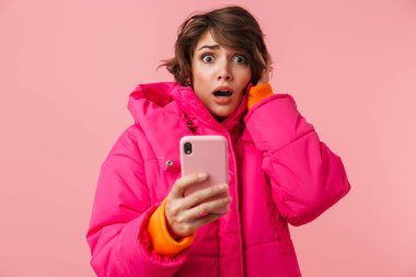 Pembe arka planda izole edilmiş cep telefonu kullanan, korkmuş genç bir kadının portresi.