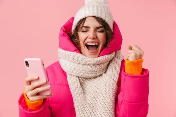 Pembe arka planda izole edilmiş, kalın elbiseli, çığlık atan ve cep telefonu kullanan heyecanlı genç bir kadının portresi.