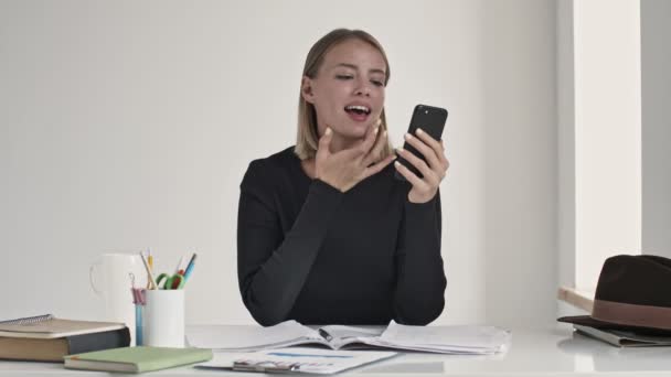 一位年轻貌美的金发女士坐在室内的一个白色办公室里 一边摸着自己的脸 一边看她的智能手机 — 图库视频影像