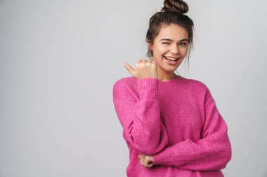 Pembe kazaklı neşeli bir genç kadın. Parmakla gösterip gri arka planda izole edilmiş bir şekilde göz kırpıyor.