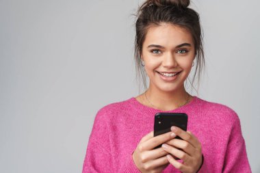 Gri arka planda mutlu, güzel bir kadının akıllı telefonuyla yazması ve gülümsemesi.