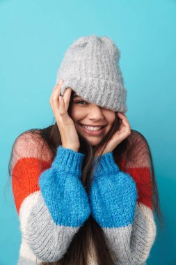 Gülümseyen genç bir kadının fotoğrafı. Mavi duvar arkasında izole bir şekilde poz veriyor. Şapkayla yüzünü örtüyor..