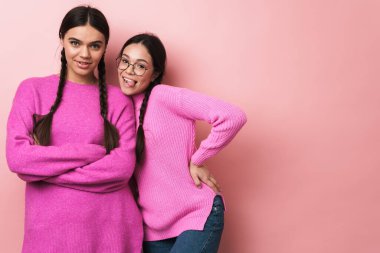Pembe arka planda izole edilmiş, günlük kıyafetleriyle kameraya gülümseyen iki mutlu genç kızın görüntüsü.