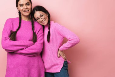Pembe arka planda izole edilmiş, günlük kıyafetleriyle kameraya gülümseyen iki mutlu genç kızın görüntüsü.