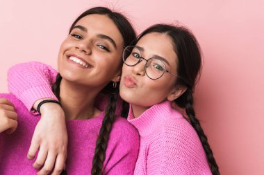 Pembe arka planda izole edilmiş, günlük kıyafetleriyle kameraya gülümseyen iki mutlu genç kızın görüntüsü.