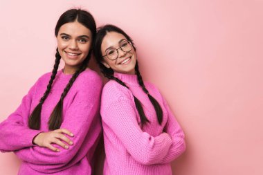Pembe arka planda izole edilmiş, günlük kıyafetleriyle kameraya gülümseyen iki mutlu genç kızın görüntüsü.