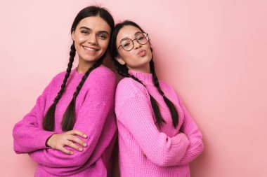 Pembe arka planda sırt sırta duran iki neşeli genç kız.