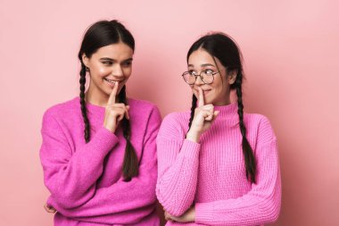 Örgülü, parmaklarını dudaklarında tutan ve pembe arka planda sessizce izole olmak isteyen iki genç kız.