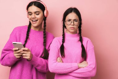 Pembe arka planda cep telefonu olan mutlu kız arkadaşının yanında duran üzgün genç kız görüntüsü.