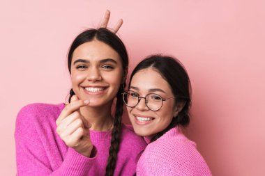 Pembe arka planda izole edilmiş, günlük kıyafetleriyle kameraya gülümseyen iki mutlu genç kızın görüntüsü.
