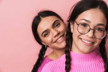 İki neşeli genç kız pembe arka planda izole edilmiş, somurtuyor, kameraya bakıyor.