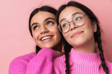 Pembe arka planda izole edilmiş, günlük kıyafetleriyle kameraya gülümseyen iki mutlu genç kızın görüntüsü.