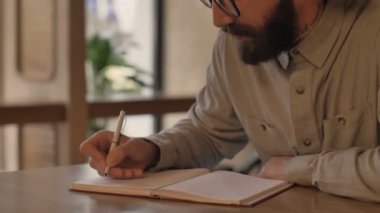Yakışıklı, konsantre sakallı adam kafeteryada deftere notlar yazıyor.