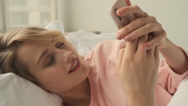 금발의 침대에서 전화를 사용하여 집안에서 행복하게 웃는다 — 비디오