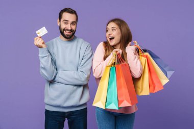 Heyecanlı, mutlu, iyimser bir çiftin resmi. Mor duvar arkasında izole edilmiş, ellerinde alışveriş çantaları ve kredi kartı var..