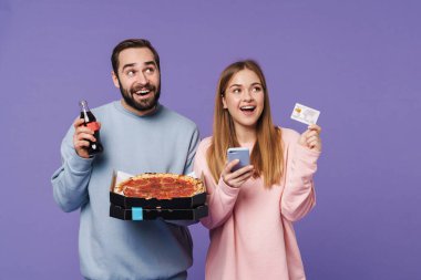 Mor duvar arkasında izole edilmiş, gazoz içen, elinde cep telefonu ile pizza yiyen mutlu bir çift görüntüsü..