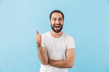 Mavi arka planda tek başına duran, mutlu, heyecanlı, sakallı ve tişört giyen genç bir adamın portresi.