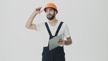 Düşünceli, genç sakallı bir inşaatçı, beyaz duvar arkasında, kask takmış, not defterine notlar yazıyor..