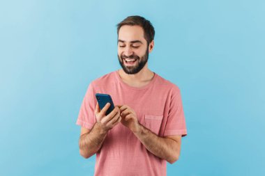 Mavi arka planda izole edilmiş, cep telefonu kullanan ve tişört giyen neşeli sakallı genç bir adamın portresi.