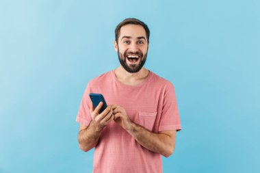 Mavi arka planda izole edilmiş, cep telefonu kullanan ve tişört giyen neşeli sakallı genç bir adamın portresi.