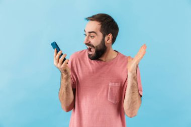 Genç, yakışıklı, şaşırmış, pozitif adamın görüntüsü mavi duvar arka planında akıllı telefon kullanılarak izole edilmiş..