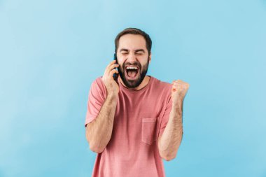 Mavi duvar arkasında izole edilmiş, cep telefonuyla konuşan genç, yakışıklı, şaşırmış bir adamın görüntüsü..