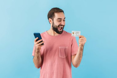 Mavi arka planda izole edilmiş, cep telefonu kullanan, kredi kartını gösteren, alışveriş yapan, tişört giyen neşeli sakallı genç bir adamın portresi.