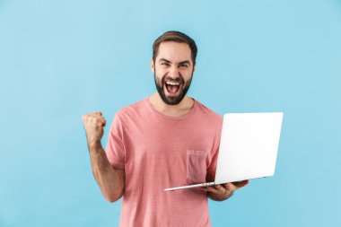 Mavi arka planda izole bir şekilde duran, dizüstü bilgisayarı tutan, başarıyı kutlayan, mutlu, sakallı, genç bir adamın portresi.