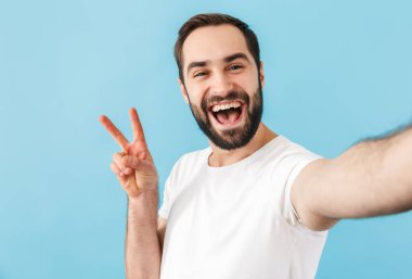 Mavi duvar arkasında izole edilmiş, heyecanlı, neşeli genç bir adamın görüntüsü barış jesti gösteren bir selfie çekin..