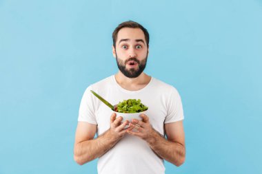 Tişört giyen yakışıklı genç sakallı adam mavi arka planda tek başına duruyor, elinde taze salata kasesi var.