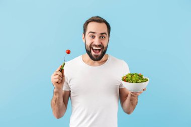 Yakışıklı, sakallı, tişört giyen, mavi arka planda tek başına duran, elinde taze salatayla kaseyle yemek yiyen genç bir adam.