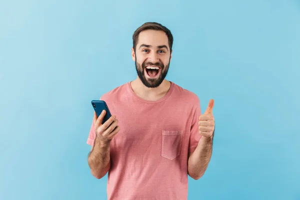 一个喜怒无常的年轻大胡子男子的画像 身穿T恤 孤零零地站在蓝色背景下 用手机 竖起大拇指 — 图库照片