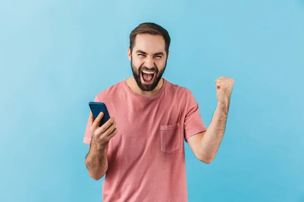 青の背景に隔離されたTシャツを着た若い陽気な興奮した髭の男の肖像画 携帯電話を使用して 成功を祝う — ストック写真