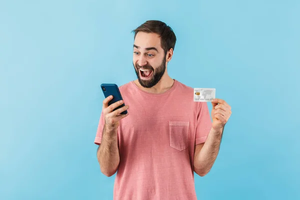 一个喜怒无常的年轻大胡子男子的画像 身穿T恤衫 孤零零地站在蓝色背景下 用手机 展示信用卡 — 图库照片