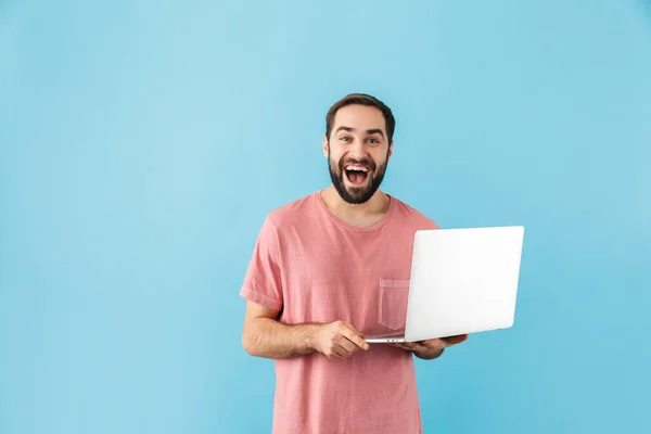 一个喜怒无常的年轻大胡子男子穿着T恤 孤零零地站在蓝色背景下 手持笔记本电脑的画像 — 图库照片