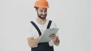 Neşeli, genç, sakallı bir inşaatçı beyaz duvar arkasında kask takarak not tutuyor..