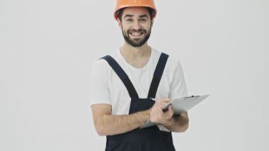 Neşeli, sakallı, genç bir inşaatçı beyaz duvar arkasına izole edilmiş, kask takmış, panoya notlar yazıyor..