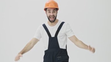 Gülümseyen, sakallı, genç bir inşaatçı beyaz duvar arkasında kendini gösteren kaskla izole edilmiş.